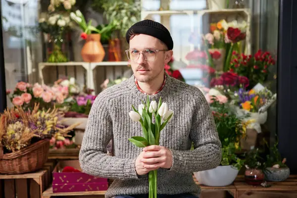 Attractive young caucasian man wearing eyeglasses and beanie in flower store with tulips bouquet. Good looking male florist in sweater holding bouquet. Small business concept. High quality image
