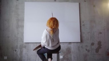 Female painter sitting in front of big white canvas holding paintbrushes and palette. Caucasian redhead artist waiting for inspiration meditating in front of blank canvas. High quality 4k footage