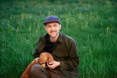 Cute male in baseball hat sitting on a grass in park and playing with small brown dachshund puppy. Pets as family members concept image. High quality image
