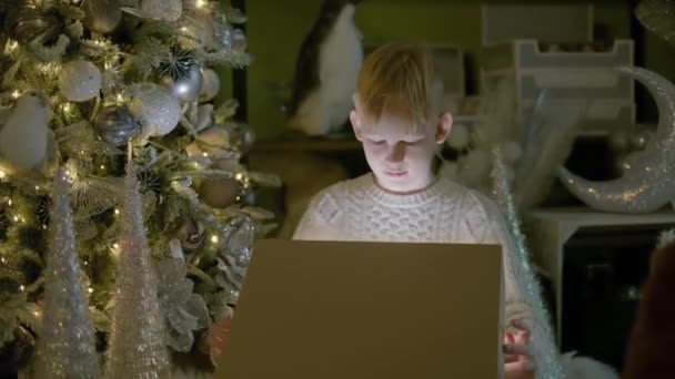 家の大きな箱に新年の贈り物をアンパッククリスマスツリーの装飾の近くに座ってセーターを着て幸せな笑顔の赤毛の子供の男の子 高品質4Kビデオ映像 — ストック動画