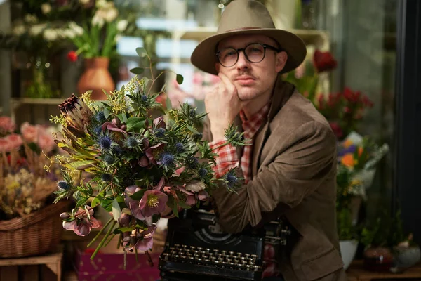 Cute caucasian male person in eyeglasses and hat sitting with old typewriter in flower shop. Poet or writer working concept. High quality photo