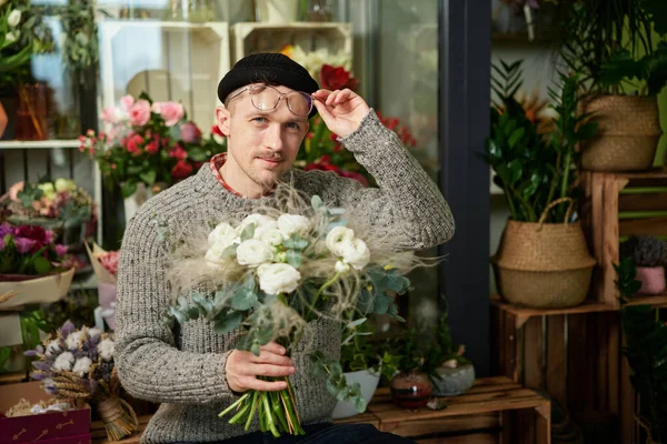 Good looking caucasian florist man wearing sweater, beanie and eyeglasses holding flower bouquet. Small business concept. Bearded male with bouquet of white roses in flower store. High quality image