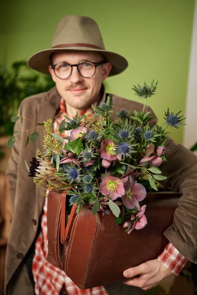 Romantic male person in eyeglasses and hat staying with flower bouquet and looking in camera in flower shop. Poet or writer portrait. High quality vertical image
