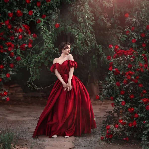 Bella donna in abito rosso passeggiando nel giardino pieno di rose. — Foto Stock