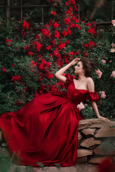 Gyönyörű, vörös ruhás nő, aki a rózsákkal teli kertben sétál.. — Stock Fotó