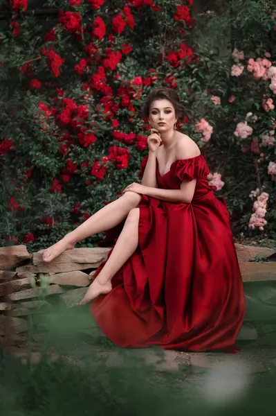 Όμορφη γυναίκα με κόκκινο φόρεμα περπατώντας στον κήπο γεμάτη τριαντάφυλλα. — Φωτογραφία Αρχείου