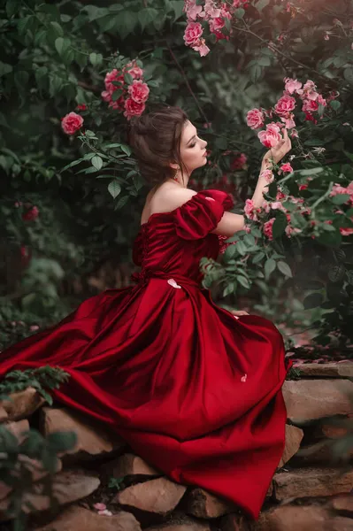 Schöne Frau in rotem Kleid spaziert durch den Garten voller Rosen. — Stockfoto