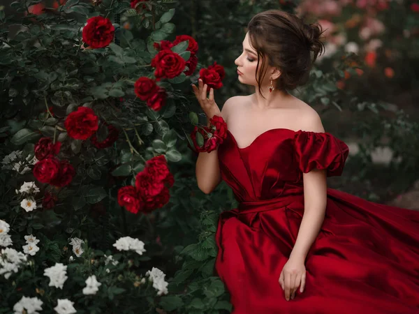 Hermosa mujer en vestido rojo caminando en el jardín lleno de rosas. — Foto de Stock
