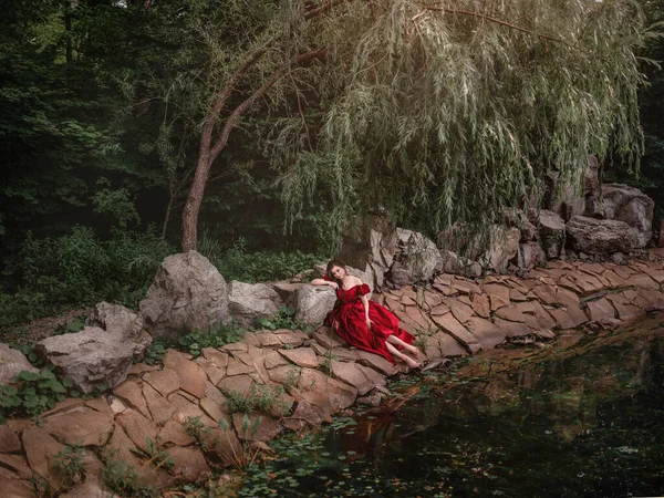 Vacker kvinna i röd klänning går i trädgården full av rosor. — Stockfoto