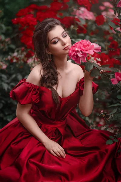 Mooie vrouw in rode jurk wandelen in de tuin vol rozen. — Stockfoto