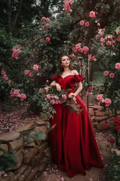 Bella donna in abito rosso passeggiando nel giardino pieno di rose. — Foto Stock