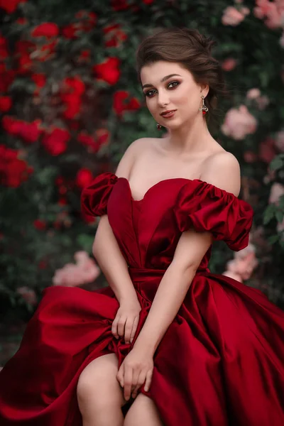 빨간 드레스를 입은 아름다운 여인 이 장미가 가득 한 정원을 걷고 있다. — 스톡 사진
