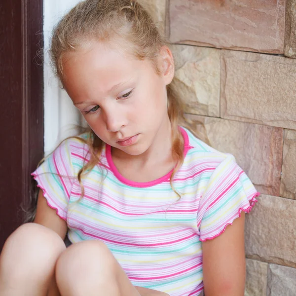 Trauriges kleines Mädchen, das neben einer Tür auf dem Hintergrund eines Marktes sitzt — Stockfoto