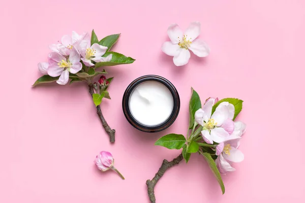 Spring concept of natural organic cosmetics. Clean brown glass cosmetic bottles with cream, delicate spring flowers on pink background flat lay top view. Beauty herbal product spa aroma oil.