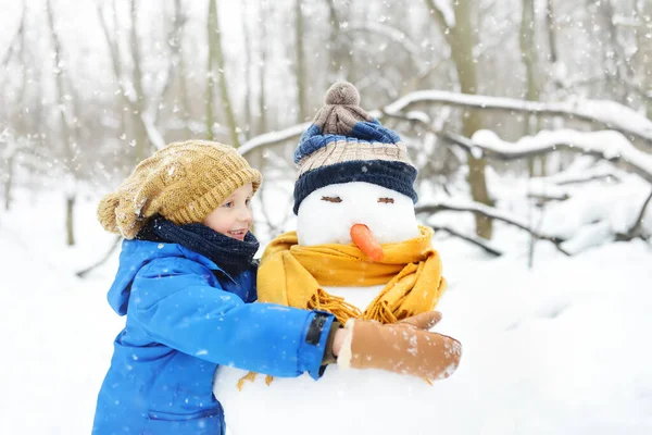 雪の公園に雪だるまを作る小さな男の子 帽子とスカーフを身に着けている雪だるまを受け入れる子供 冬に子供連れの家族とのアクティブな屋外レジャー 雪の冬の公園を散歩中の子供 — ストック写真