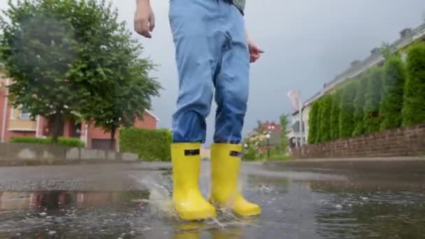 小さな町で雨の夏の日に水の水たまりにジャンプ黄色のゴムブーツを着て小さな男の子のスローモーションビデオ 子供は楽しんでる 雨の中で子供のための屋外ゲーム — ストック動画