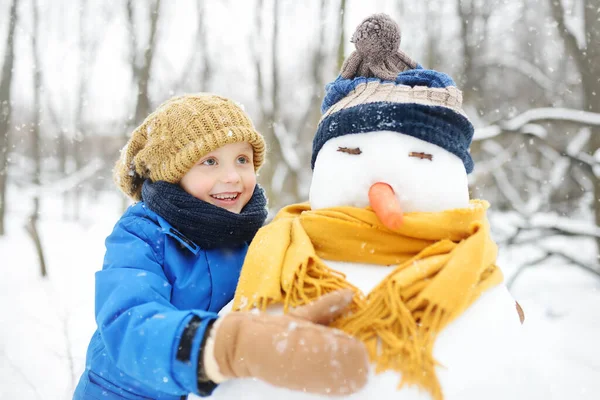 雪の公園に雪だるまを作る小さな男の子 帽子とスカーフを身に着けている雪だるまを受け入れる子供 冬に子供連れの家族とのアクティブな屋外レジャー 雪の冬の公園を散歩中の子供 — ストック写真