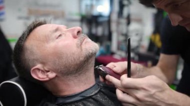 Kuaförde tıraş makinesi kullanan yakışıklı, olgun sakallı bir adam. Saç sanatçısı, erkek berber dükkanında sakal yapıyor. Profesyonel stilistin hizmetleri. Erkekler için moda saç bakımı