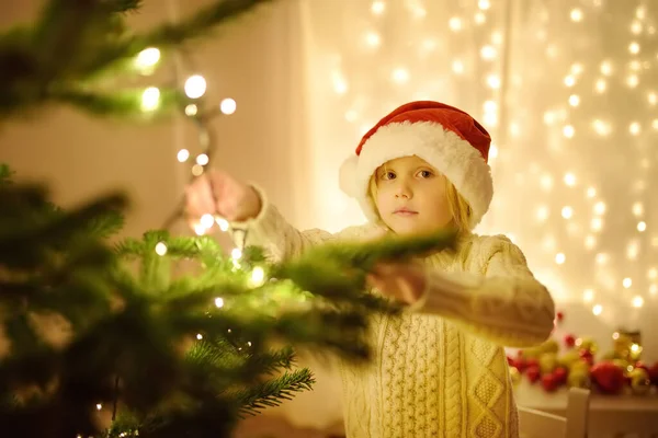 サンタの帽子をかぶっている小さな男の子はクリスマスを祝う準備ができている ガーランドでクリスマスツリーを飾るかわいい子供 新年の夜に魔法と贈り物の赤ちゃんの希望 冬の休日の周りの伝統 — ストック写真