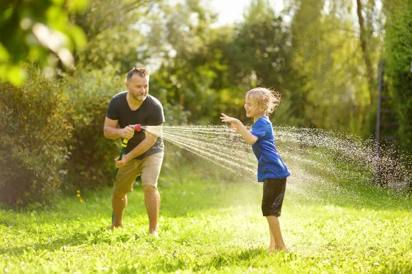 日当たりの良い裏庭で庭のホースで遊んで彼の父と面白い小さな男の子 水のスプレーで楽しんでいる未就学児 子供連れの家族のための夏の屋外活動 — ストック写真