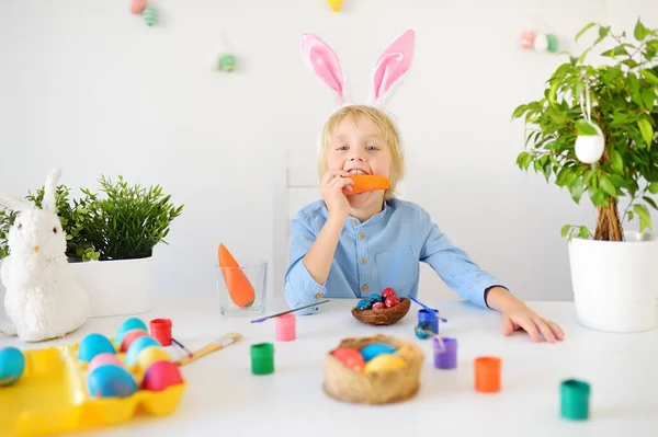 Cute playful boy is wearing bunny ears paints eggs for Easter holiday. Child is eating of carrot as bunny. Preparing for the holiday is a joy for kids. Celebration Easter at home.