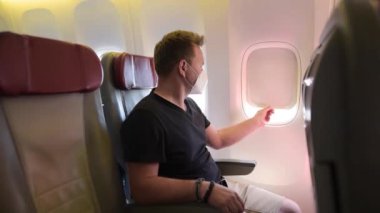 Uçakla seyahat eden maskeli orta yaşlı bir adam. Uçakta oturan ve uçuş sırasında uçak camına bakan yolcu. Koronavirüs salgını sırasında sinek taşımacılığının güvenliği ve korunması