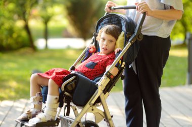 Yazın parkta tekerlekli sandalyeli engelli bir kızla yürüyen bir kadın. Bebek bacaklarının ortozu. Çocuk serebral felci. Karargahın tedavisi ve rehabilitasyonu. Engelli çocuğu olan bir aile..