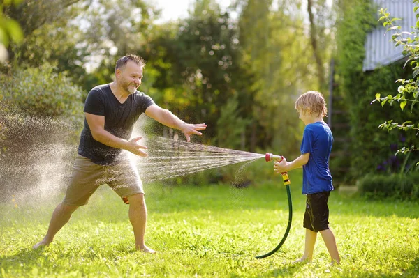 日当たりの良い裏庭で庭のホースで遊んで彼の父と面白い小さな男の子 水のスプレーで楽しんでいる未就学児 子供連れの家族のための夏の屋外活動 — ストック写真