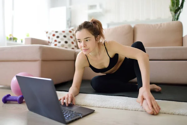 远距离运动健身训练 一个有魅力的年轻女人在家用笔记本电脑做瑜伽或在网上伸展身体 通过因特网进行的娱乐 教育和体育活动 — 图库照片