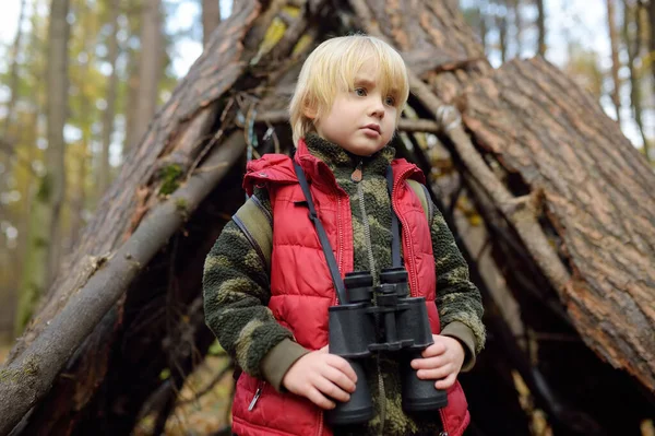 在秋天的森林里徒步旅行时 小男孩用双筒望远镜侦察 孩子的后面是小茅屋 有子女家庭探险 探险和远足旅游的概念 — 图库照片