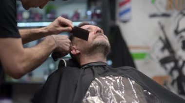Kuaförde tıraş makinesi kullanan yakışıklı, olgun sakallı bir adam. Saç sanatçısı, erkek berber dükkanında sakal yapıyor. Profesyonel stilistin hizmetleri. Moda saç bakımı 