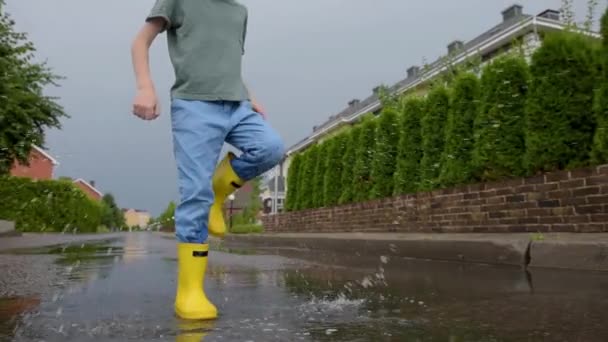小さな町で雨の夏の日に水の水たまりにジャンプ黄色のゴムブーツを着て小さな男の子 子供は楽しんでる 雨の中で子供のための屋外ゲーム — ストック動画