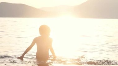 Mutlu çocuk gün batımında denizde yüzer. Neşeli çocuk gölde yüzerken su sıçrıyor ve eğleniyor. Okul tatilinde çocuklar için aktif boş zaman