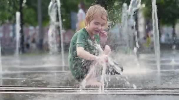 晴れた夏の日には噴水のウォータージェットの間の広場で小さな男の子が遊んでいます 大都市の子供たちのためのアクティブな夏のレジャー — ストック動画