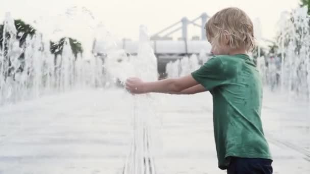晴れた夏の日には噴水のウォータージェットの間の広場で小さな男の子が遊んでいます 大都市の子供たちのためのアクティブな夏のレジャー — ストック動画
