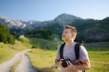Mature man tourist with backpack and camera is hiking on mountain valley. Photographer taking a picture during travel. Tourism and adventure.