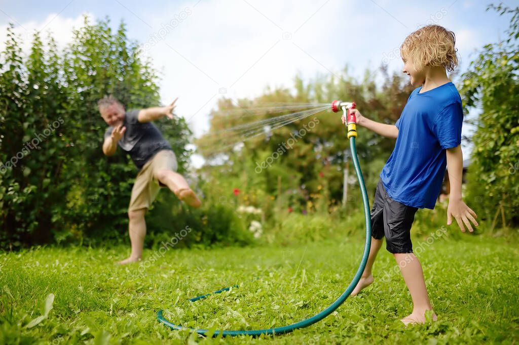 Funny little boy with his father playing with garden hose in sunny backyard. Preschooler child having fun with spray of water. Summer outdoors activity for family with kids. Dad involved in parenting