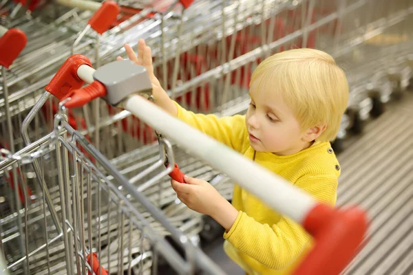A cute blonde boy plays with shopping carts at the entrance to the supermarket. The curious child explore how the world around works. Going to the grocery store for family with kids