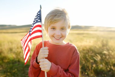 Cute little boy celebrating of July, 4 Independence Day of USA at sunny summer sunset. Child running and jumping with american flag symbol of United States over wheat field. Pride and patriotism