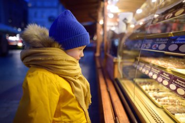 Little boy boy chooses sweets (fruit in chocolate glaze) on street Christmas market. Famous fruits, candy, cookies, gingerbread in chocolate glaze on skewers in bakery Xmas Eve. Christmas european food.
