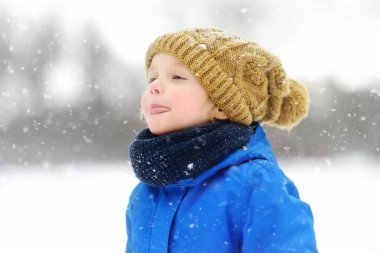Sıcak bir şapka takan tatlı çocuk diliyle kar taneleri yakalıyor. Mavi kışlık elbiseli komik küçük çocuk kar yağışı sırasında yürür. Çocuklar için açık hava kış aktiviteleri.