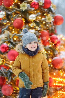 Küçük çocuk, altın, kırmızı toplar ve çiçeklerle süslenmiş yapay Noel ağacına hayran. Geleneksel büyük şehir noel pazarında. Şehir dekorasyonu kutluyor. İyi tatiller..