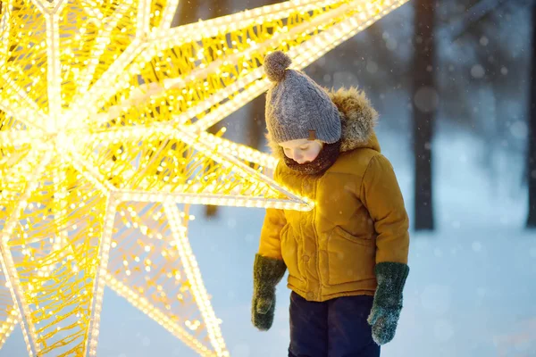 小さな男の子はクリスマススターの形で大きな輝く通りの装飾を賞賛しています オープンエアの伝統的な都市のクリスマスマーケット 現代の都市のお祝いの装飾 幸せな休日 — ストック写真