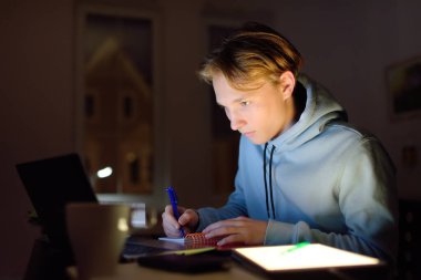 Yoğunlaşmış genç, gece geç saatlere kadar ofiste bilgisayar ve tabletle ders çalışıyor. Uzaktan çalışma, uzaklık öğrenme, ek eğitim, serbest çalışma teknolojileri