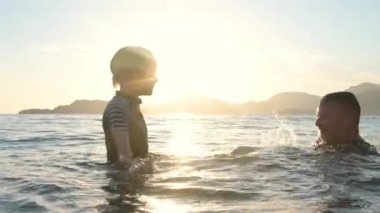 Mutlu çocuk ve olgun babası gün batımında denizde yüzerken birlikte eğleniyorlar. Neşeli çocuk ve babası gölde yüzerken su sıçratıyor. Tatillerde aile için aktif eğlence.