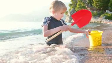 Deniz kıyısında oturan kumla ve taşlarla oynayan mutlu çocuk. Neşeli çocuk açık deniz kıyısında bir plaj tatili sırasında eğleniyor. Okul tatilinde çocuklar ve tüm aile için boş zaman.