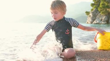 Deniz kıyısında oturan kumla ve taşlarla oynayan mutlu çocuk. Neşeli çocuk açık deniz kıyısında bir plaj tatili sırasında eğleniyor. Okul tatilinde çocuklar ve tüm aile için boş zaman.