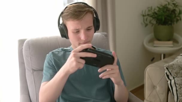 十几岁的男孩在玩一个便携式游戏机 一个青少年坐在客厅的椅子上玩小玩意 博彩青年 — 图库视频影像