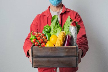 Koruyucu maskeli genç teslimatçı elinde tahta kutu tutuyor. Alışveriş kutusu olan bir kurye. Süpermarketten eve güvenli teslimat. COVID-19 koronavirüs koruması.