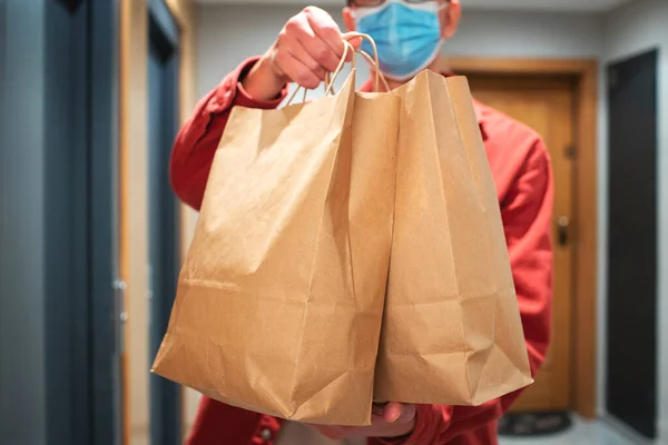 Delivery man in protective mask holding paper bag with food in the entrance. The courier gives the box with fresh vegetables and fruits to the customer.